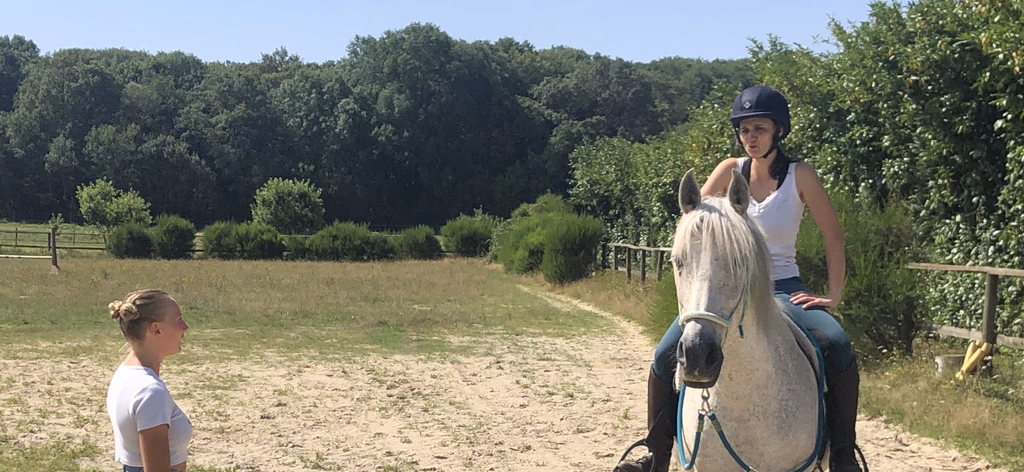 Cours d'équitation en carrière : cavalière sur un cheval gris guidée par son enseignante